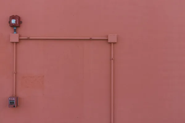 Surface-mounted electrical conduit and a red fire alarm running across a bare salmon-pink exterior wall.