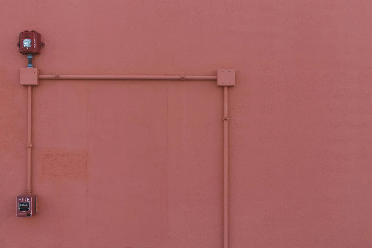 Surface-mounted electrical conduit and a red fire alarm running across a bare salmon-pink exterior wall.
