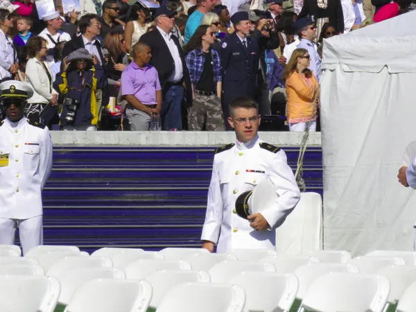 A newly commissioned naval officer in dress whites holds his cover to his chest, standing among rows of empty white chairs as a crowd looks on behind him.