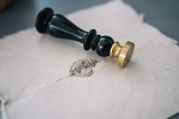 A black wax seal stamp with a brass die resting on handmade paper beside its pressed silver impression.