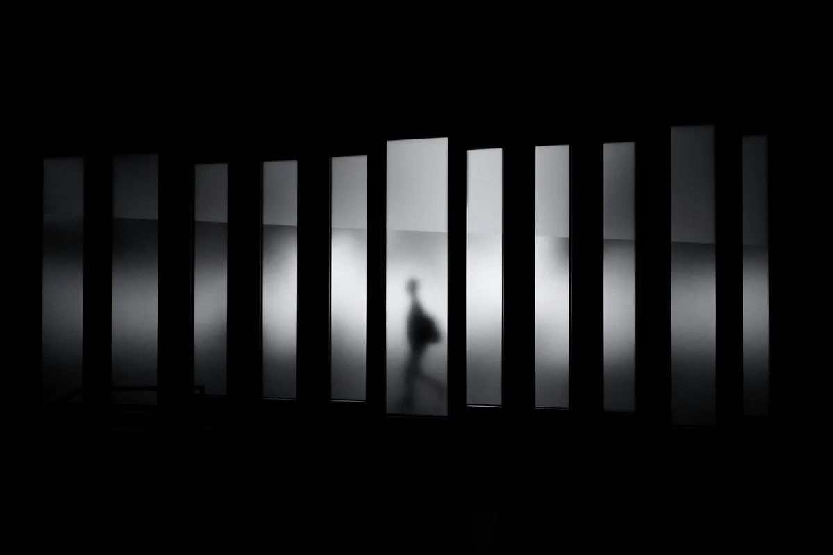 A high-contrast silhouette of a figure walking through a corridor of vertical light and shadow.