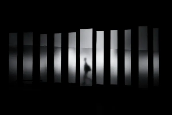 A high-contrast silhouette of a figure walking through a corridor of vertical light and shadow.