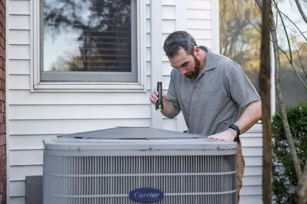 A technician inspects a home air-conditioning unit by flashlight, the diagnostic moment behind a repair quote.